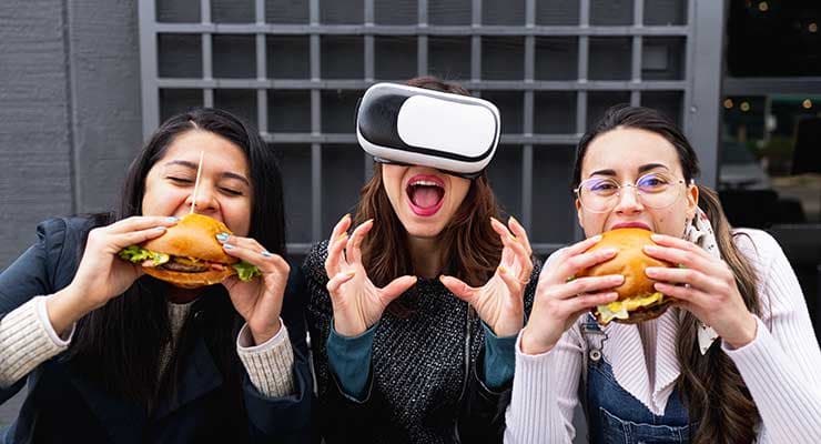 2 mujeres comen hamburguesas reales, una mujer con gafas VR